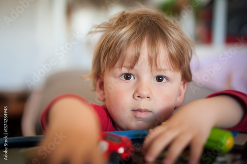 A young boy with tousled hair sits in a cozy living room, reaching for colorful toys laid out before him. His intense gaze shows concentration and curiosity as he plays