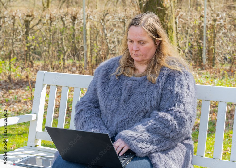 Frau in einer Kaninchenfell Jacke arbeitet im Garten mit einem Notebook im Homeoffice  
