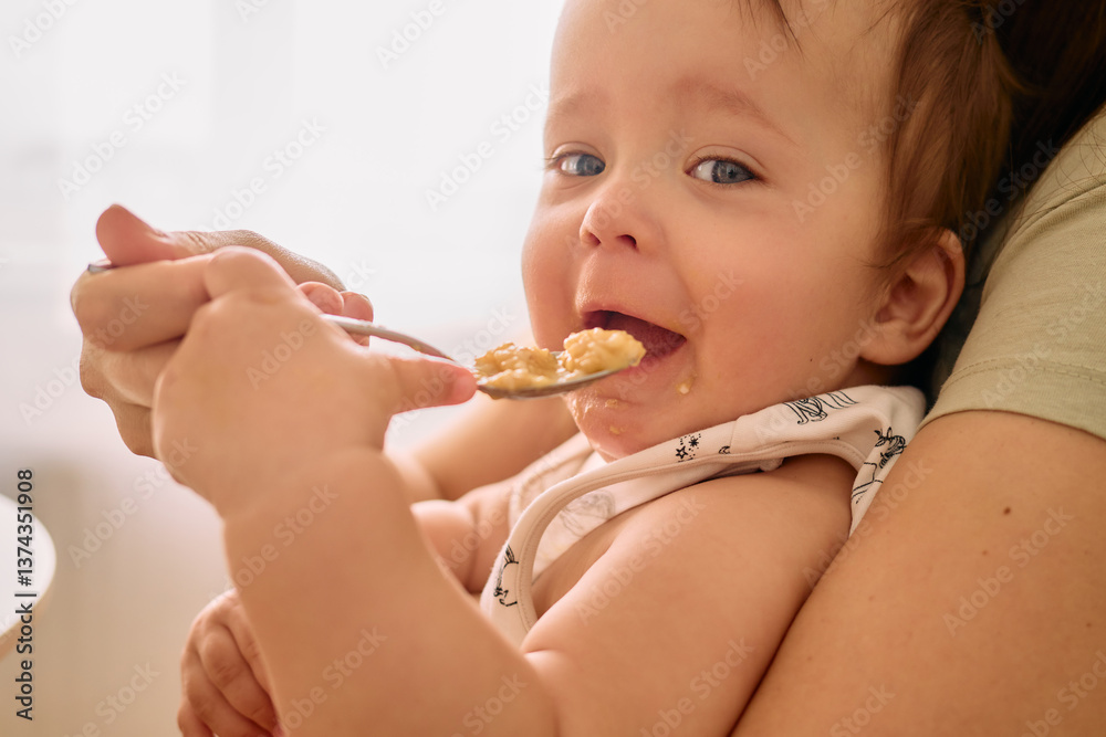 A joyful baby with blue eyes smiles while being fed oatmeal by a caregiver. Soft light fills the cozy kitchen, creating a warm atmosphere during breakfast time A joyful baby with blue eyes smiles while being fed oatmeal by a caregiver. Soft light fills the cozy kitchen, creating a warm atmosphere during breakfast time