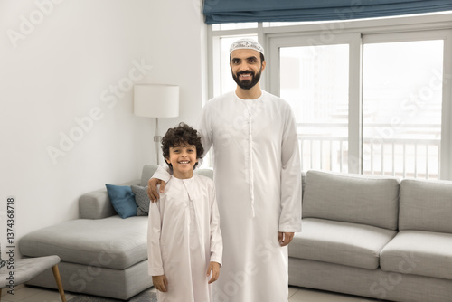 Young 30s Arabic father and little 7s son standing together in living room, both dressed in traditional white thobes, look at camera with smile. Family ties, tradition, cultural pride, paternal care