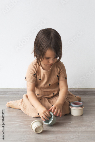 Little girl in beige dress with black dots sitting on the floor and playing with stacking cups, focused on the activity in a calm indoor setting, representing childhood, learning and play, vertical