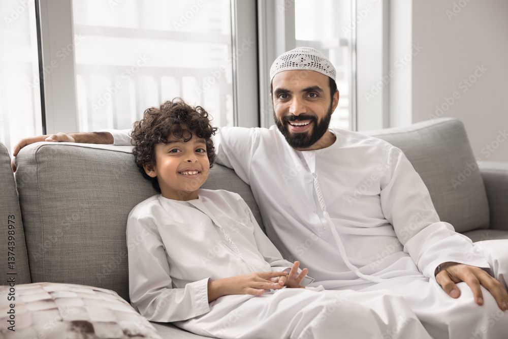 Obraz premium Joyful Arabic man his little kid sit together on couch, dressed in traditional white thobes, symbolizing cultural and familial bond. Upbringing, family unity, tradition, father-son relations, heritage