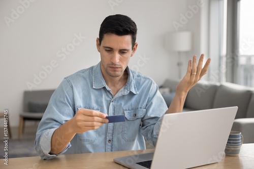 Hispanic man sit at table with laptop, holding card with concerned expression, experiences payment issue, transaction declined, feeling worried about insufficient funds, blocked card, or banking error