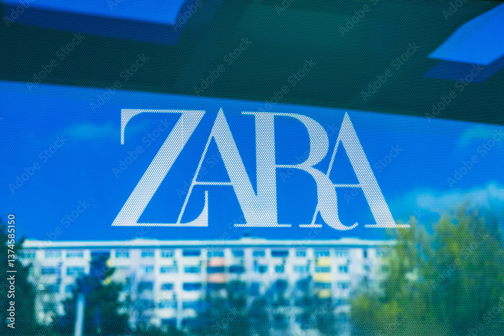 Fototapeta premium White ZARA logo on perforated glass, reflection in city street shop window in Burgas, Bulgaria, blurred urban buildings, sky in background. Fashion brand and cityscape in modern visual contrast