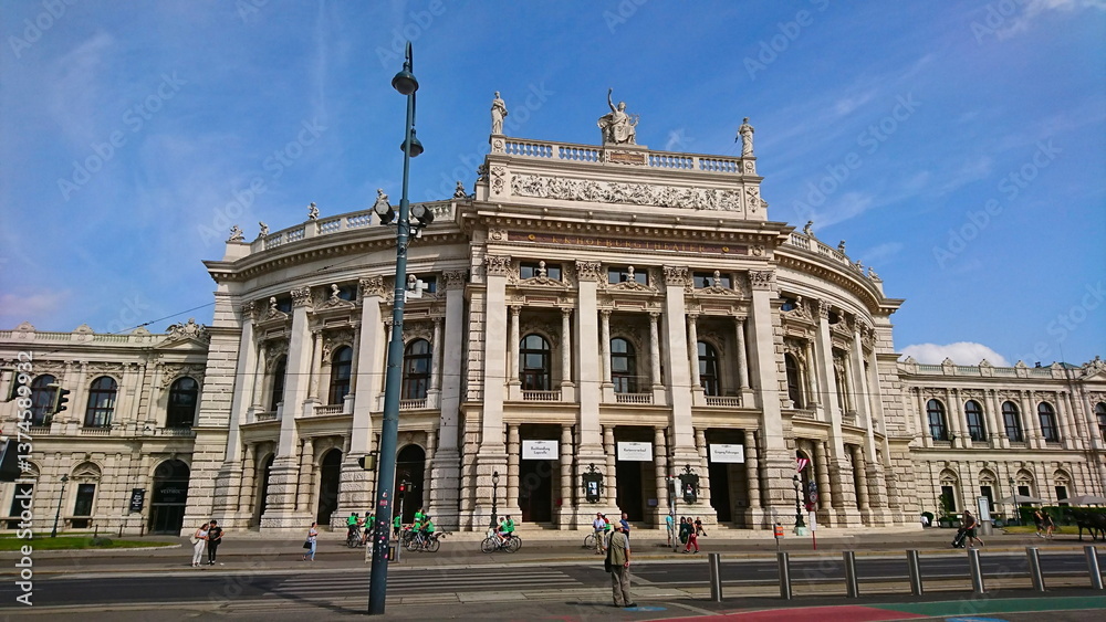 Fototapeta premium The iconic facade of the Burgtheater while cyclists and pedestrians enjoy a sunny day