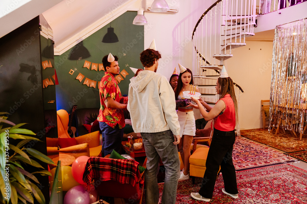 Obraz premium A group of five friends stand and look at a cake held by a female friend who walks towards them, in the living room, at a party