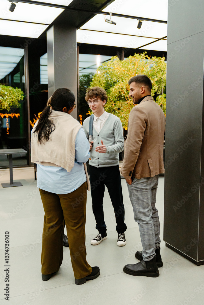 Fototapeta premium Three young colleagues of different ethnicities stand in a business hall with small trees in the background and communicate with each other. Concept of employees' informal conversation, networking.