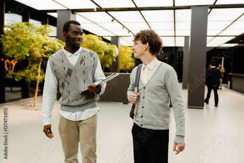 A young black man and a white man walk side by side in the modern office lobby, casually talking and holding laptops and a bag. Business context with professional attire and bright space.