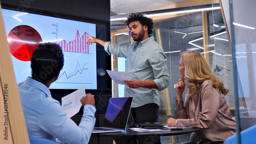 Tableau sur toile A man in a meeting explains a chart on a large screen to a group, highlighting the importance of effective communication and collaboration in a modern office setting