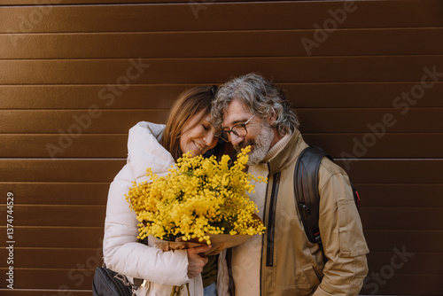 A White elder woman with brown hair, a White elder man with a beard and glasses, smiling together while holding a bouquet of yellow flowers, in a warm, sunny outdoor setting, enjoying a joyful moment.