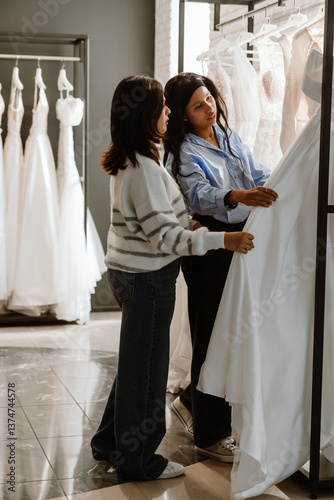 A Black bride in a blue blouse and her White female friend, shopping for wedding dresses together in a bridal store, surrounded by elegant white gowns, sharing an exciting and joyful moment.