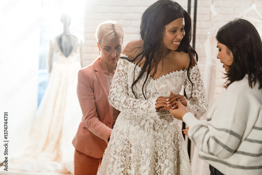 Naklejka premium A Black young bride wearing a lace wedding dress, her White young female friend, and a White adult female consultant in a bridal shop, helping with the fitting and wedding dress selection.