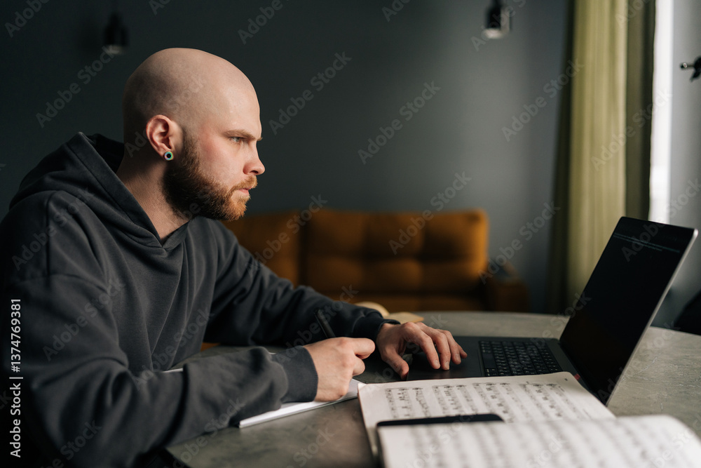Side view of talented composer writing music notes on paper sitting at table with laptop, books, sheet music in home office, immersing in creativity and inspiration. Concept of remote job, technology. Side view of talented composer writing music notes on paper sitting at table with laptop, books, sheet music in home office, immersing in creativity and inspiration. Concept of remote job, technology.