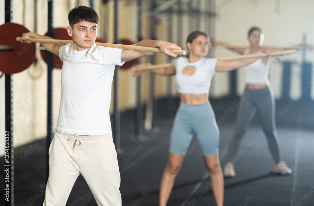Fototapeta premium Young guy, together with other athletes, does a warm-up using a long stick in the gym