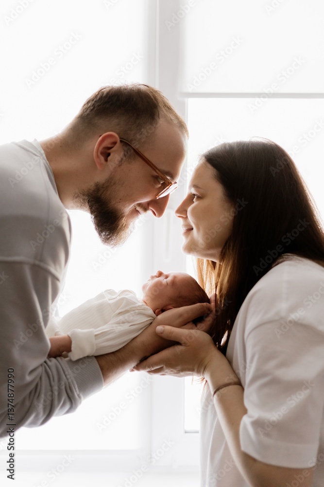 Portrait of a young family with infant in his arms photographed on a light window background. Portrait of a young family with infant in his arms photographed on a light window background.