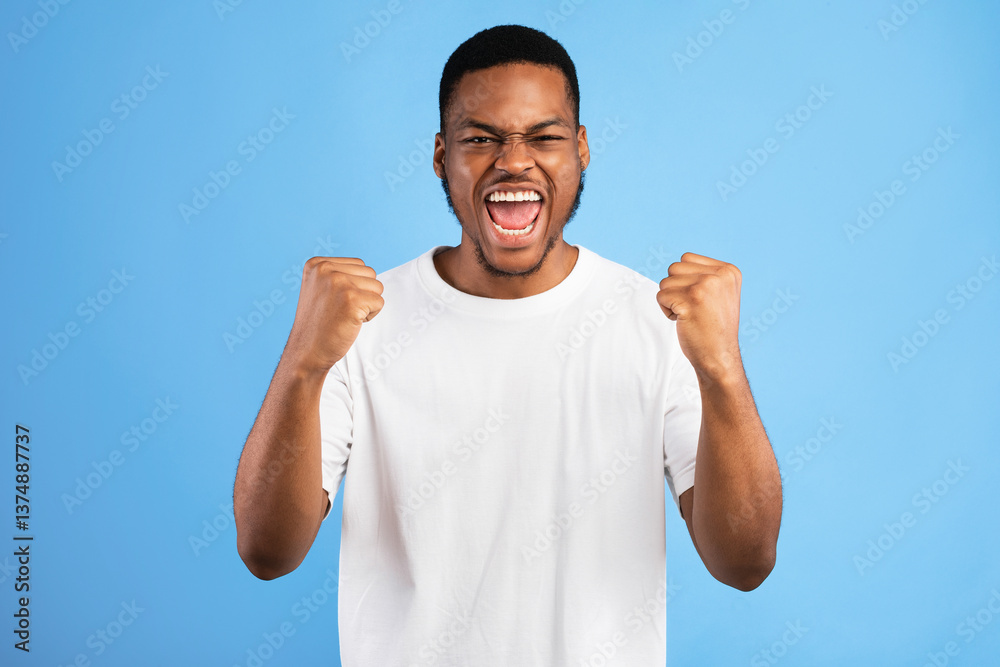 Fototapeta premium Winner. Emotional Black Guy Shouting Yes Shaking Fists Looking At Camera Standing On Blue Background. Studio Shot