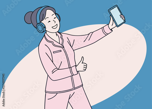 Asian woman in pink pajamas smiling with headphones around her neck, showing a thumbs up and holding a blank smartphone against a solid blue background
