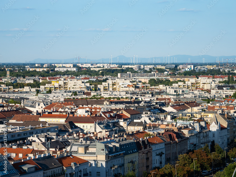 Fototapeta premium Cityscape of Vienna, Austria with a lot of residential buildings. Aerial view on the urban area of the capital city. Wind turbines are on the horizon in front of the mountains.