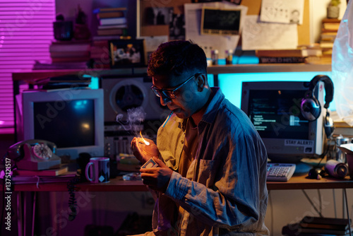 Young Indian man with glasses and hair in ponytail lighting up cigarette in dimly lit room using match