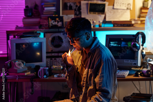Modern Indian man with glasses lighting up cigarette in dimly lit cluttered room using match