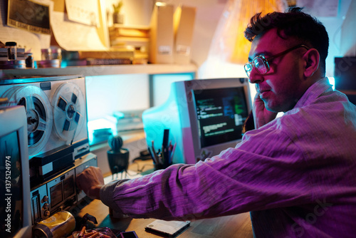 Young male hacker turning on retro tape recorder and listening to recording with headphones in room with neon light