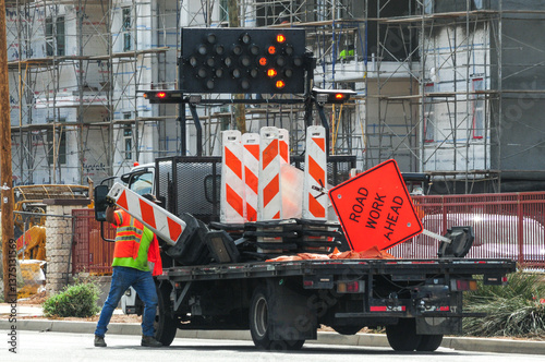 A traffic control worker unloads road barricades from a flatbed truck near a construction zone, supporting roadway safety, signage deployment, and lane management for nearby infrastructure work