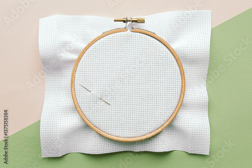 Wooden embroidery hoop with empty white canvas cloth and needle on pastel background with light pink and light green colors, ready for cross-stitching
