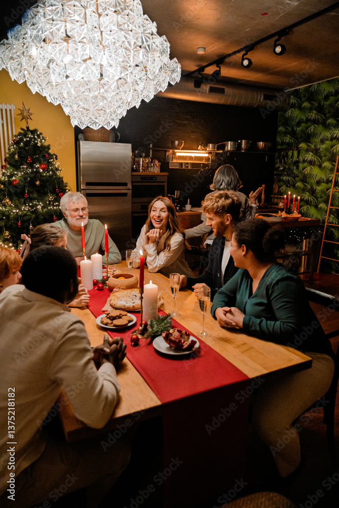 An elderly White grandfather is sitting with a multiethnic family at a festive table with food and candles and talking in a room with a Christmas tree and glass chandelier during a cozy dinner An elderly White grandfather is sitting with a multiethnic family at a festive table with food and candles and talking in a room with a Christmas tree and glass chandelier during a cozy dinner
