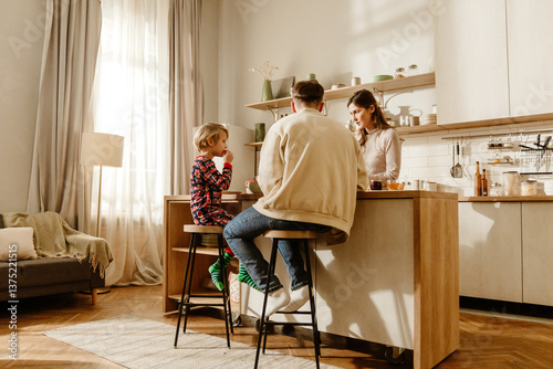 A school-aged European boy with short blond hair, having breakfast with his mid-30s parents in their modern kitchen as the dad sits next to him and the mom stands on the other side of the table