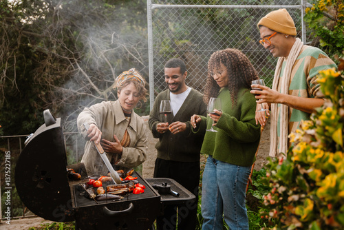 A White woman in her 30s, with a bandana around her hair, grilling some sausages and veggies on an outdoor grill in the backyard as she chats with her diverse friends who stand around her