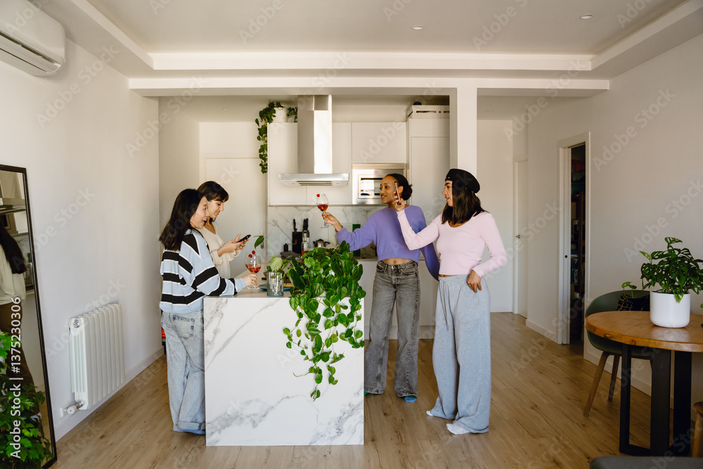 Fototapeta premium Four diverse women gather in a modern well-lit kitchen and celebrate. One holds a glass of wine while the marble-topped island holds potted plants and adds to the stylish design of the space.