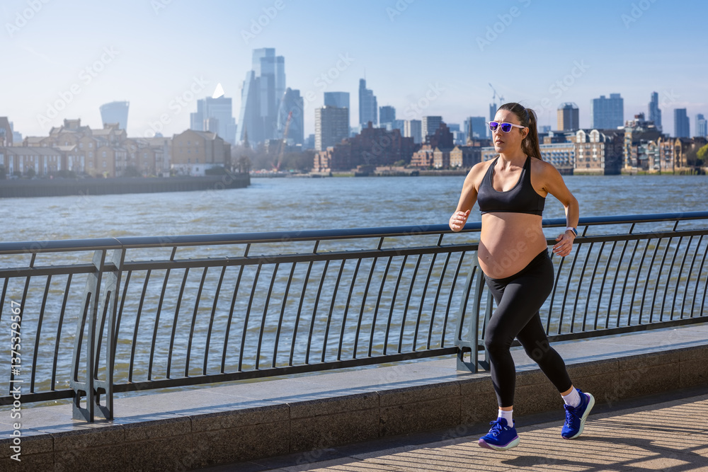 A pregnant woman in sports clothing running for exercise in front of an urban skyline during a sunny day