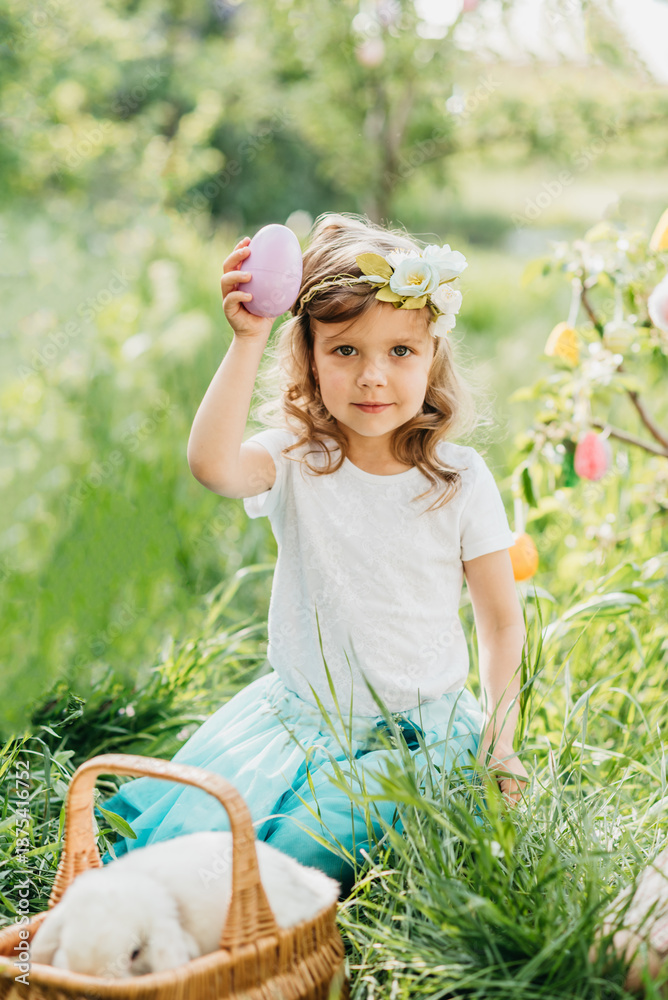 Fototapeta premium Easter egg hunt. Girl child Wearing Bunny Ears Running To Pick Up Egg In Garden. Easter tradition. Baby with basket full of colorful eggs.