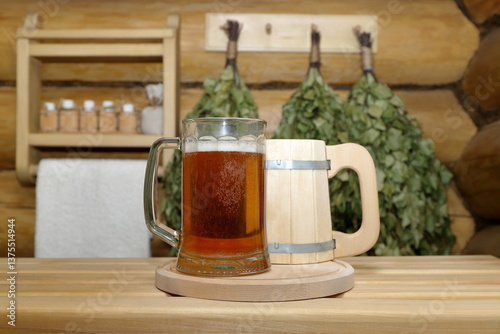In the sauna interior, on a plank bench a glass mug of light beer stands next to a wooden mug on the background of a log wall with dry birch brooms and shelf with bottles of aromatic oils and towel.