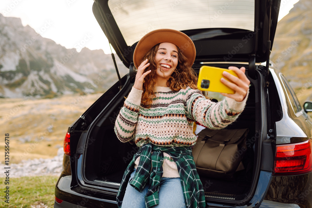 Naklejka premium Young woman with a phone enjoying the mountain landscape while sitting in the trunk of a car. Beautiful traveler blogging on a car adventure. Travel, freedom concept. Active lifestyle.
