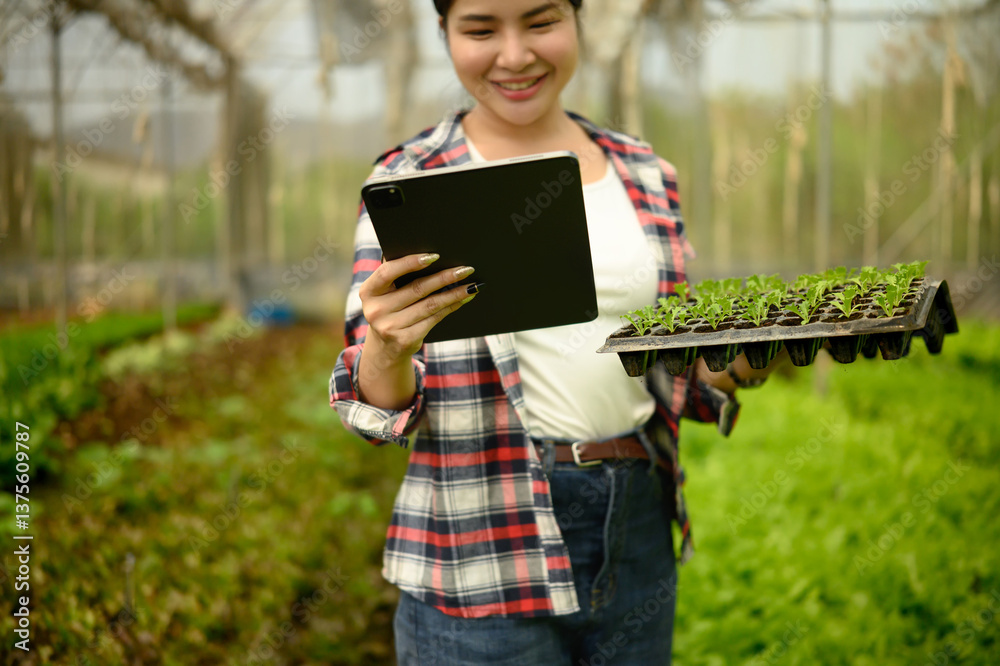 Smiling woman holding a tray of seedlings while checking data on digital tablet inside a greenhouse Smiling woman holding a tray of seedlings while checking data on digital tablet inside a greenhouse