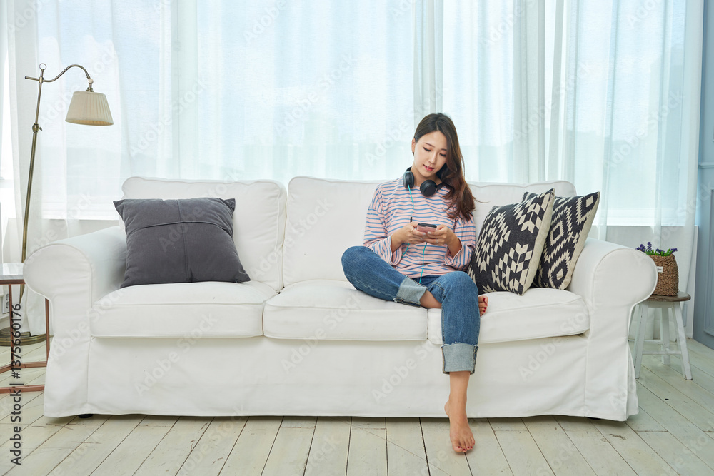 Obraz premium A young Asian woman is sitting on a living room sofa, listening to music through a headset connected to her smartphone.