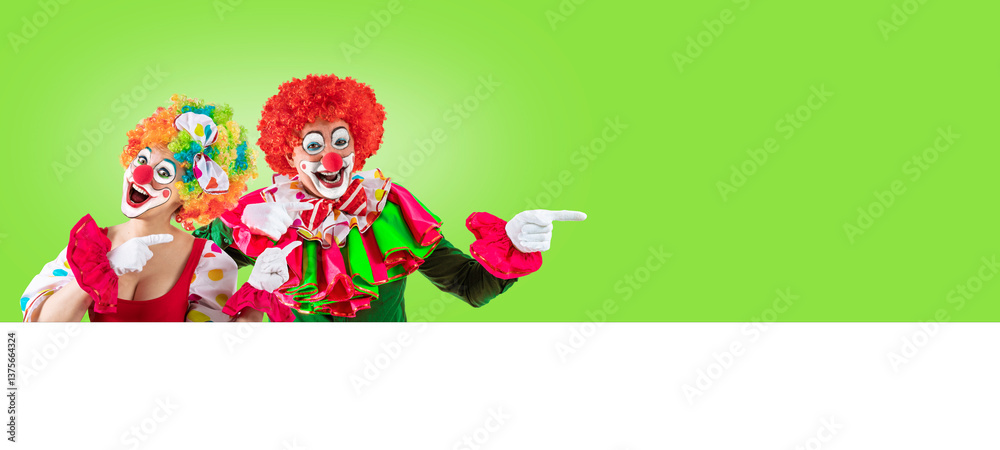 Naklejka premium Female clown and male clown pointing at blank template copy spase for advertising, posters, greetings, invitations. Entertainer as Joker in a suit and wig, with clown whiteface makeup.