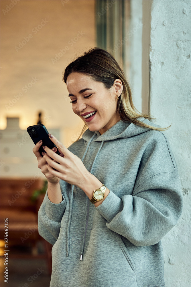 Fototapeta premium Laughing Woman Wearing Hoodie and Using Smartphone Indoors in Modern Home