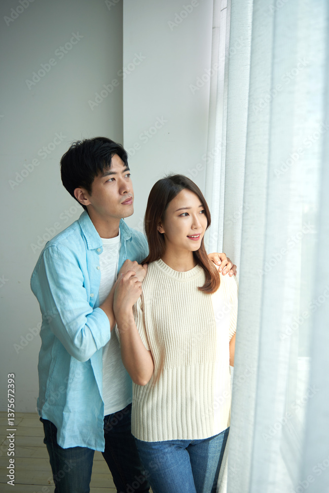 A young Asian man and a beautiful woman stand affectionately in a room with large windows decorated with white curtains. A young Asian man and a beautiful woman stand affectionately in a room with large windows decorated with white curtains.