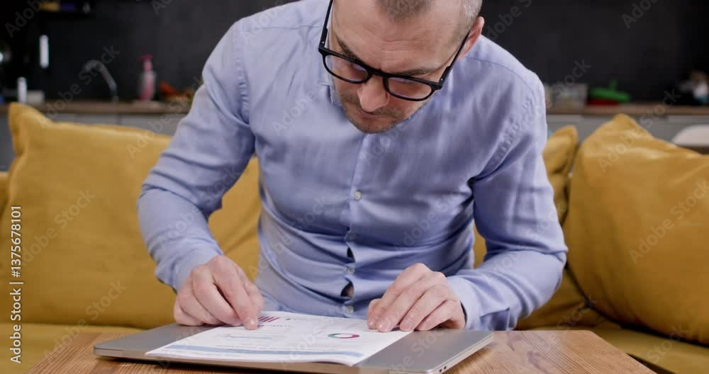 Man in glasses examines paper document with diagrams sitting on sofa. Businessman observes and calculates company data analysis in relaxed home atmosphere Man in glasses examines paper document with diagrams sitting on sofa. Businessman observes and calculates company data analysis in relaxed home atmosphere