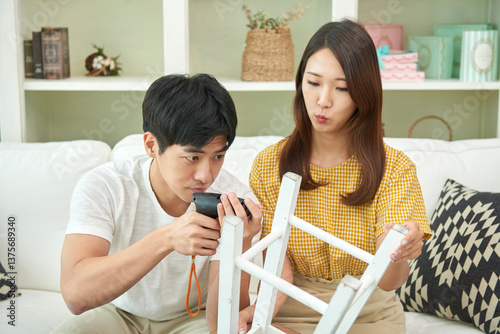 The newlywed couple sits on the living room sofa, assembling a chair with an electric drill