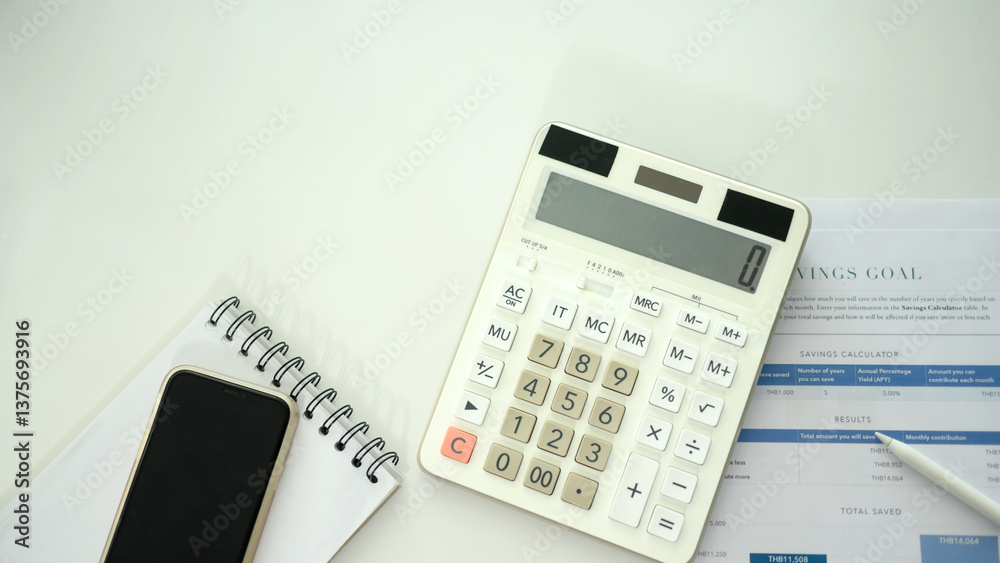 Flat lay of calculator, savings goal worksheet, notebook and smartphone on a white desk Flat lay of calculator, savings goal worksheet, notebook and smartphone on a white desk