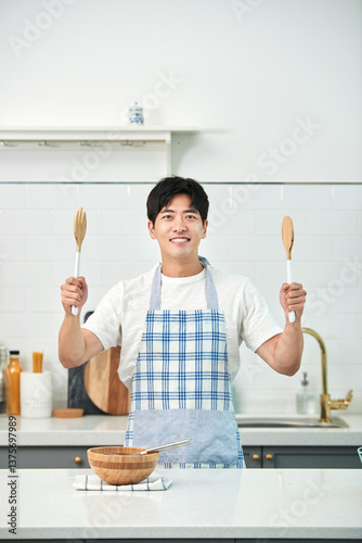 A young Asian man in the kitchen, wearing an apron, prepares a meal.