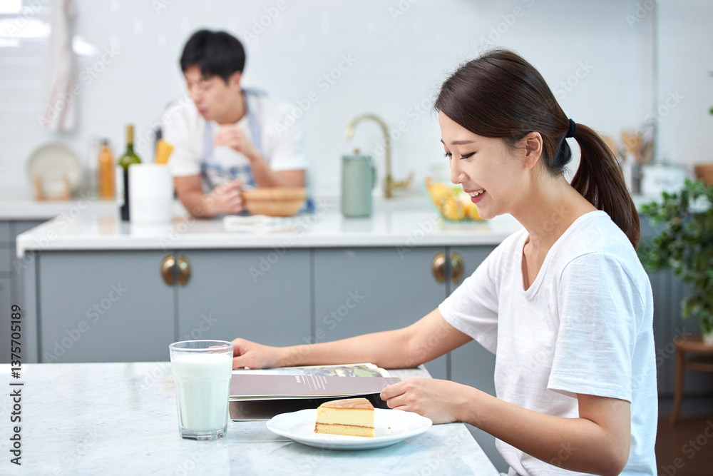 Young Asian woman in a white T-shirt sitting at a kitchen dining table chair, reading while eating bread and drinking milk Young Asian woman in a white T-shirt sitting at a kitchen dining table chair, reading while eating bread and drinking milk
