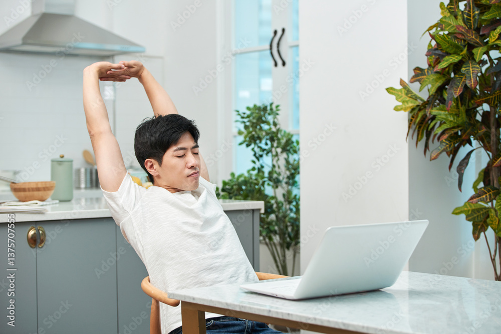 Young Asian man in a white T-shirt stretching to loosen up while using a laptop at the dining table in a living room with a kitchen Young Asian man in a white T-shirt stretching to loosen up while using a laptop at the dining table in a living room with a kitchen