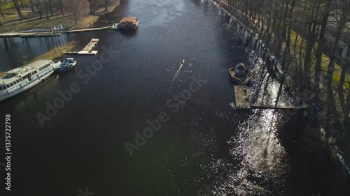 Aerial drone footage flying lower on top of Tartu cold dark colored river called Emajõgi where people are rowing a kajak or a paddle boat during spring time sunny day and large boats are achored too.