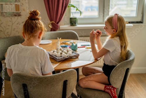two sisters, a girl of 8 years old blonde and her sister red-haired girl of 10 years old sit on a chair at home and paint a chicken white egg for the holy Orthodox holiday of bright Easter in the kitc