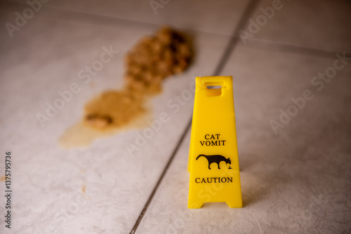 A yellow caution sign warns of cat vomit on a tiled floor. The blurred mess in the background emphasizes the humorous yet practical warning.