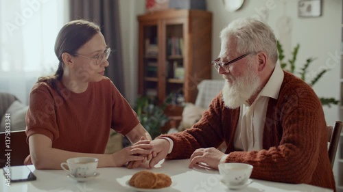 Young woman sitting at table with teacups, holding hand of elderly grandfather during heartfelt conversation, providing care and emotional support at home